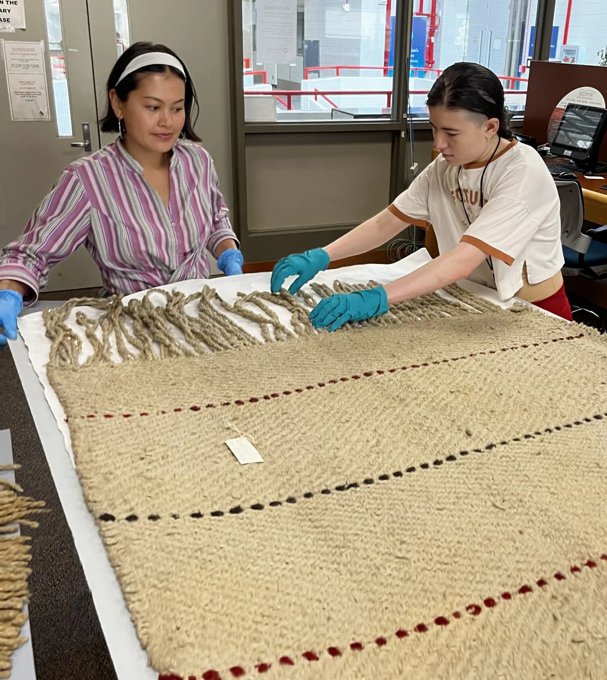 Two SIMA Collections Interns, standing at a table and looking at a large handwoven textile. Both are wearing gloves. One is straightening the fringe on the textile. 