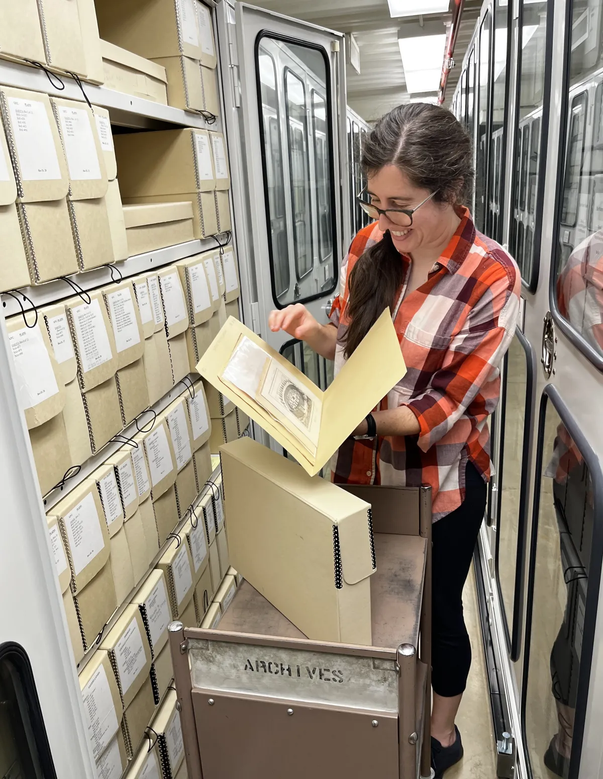 SIMA intern, standing in a long hallway lined with cabinets. She is standing in front of an open cabinet that is lined with archival boxes. One of the boxes is pulled out on a cart in front of her. She is holding a folder and looking at documents.